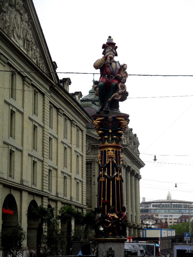 Der Kindlifresserbrunnen in der Berner Altstadt. Der Brunnen hat eine Figur, die auf einer Säule steht. Ein Mann in Rosa oder pinker Kleidung, mit einem Spitzhut auf dem Kopf. Der Mann beißt einem Baby den Kopf ab.
