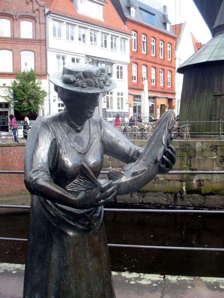 Bronzeskulptur einer Frau mit Hut, die einen Fisch in der Hand hält.
