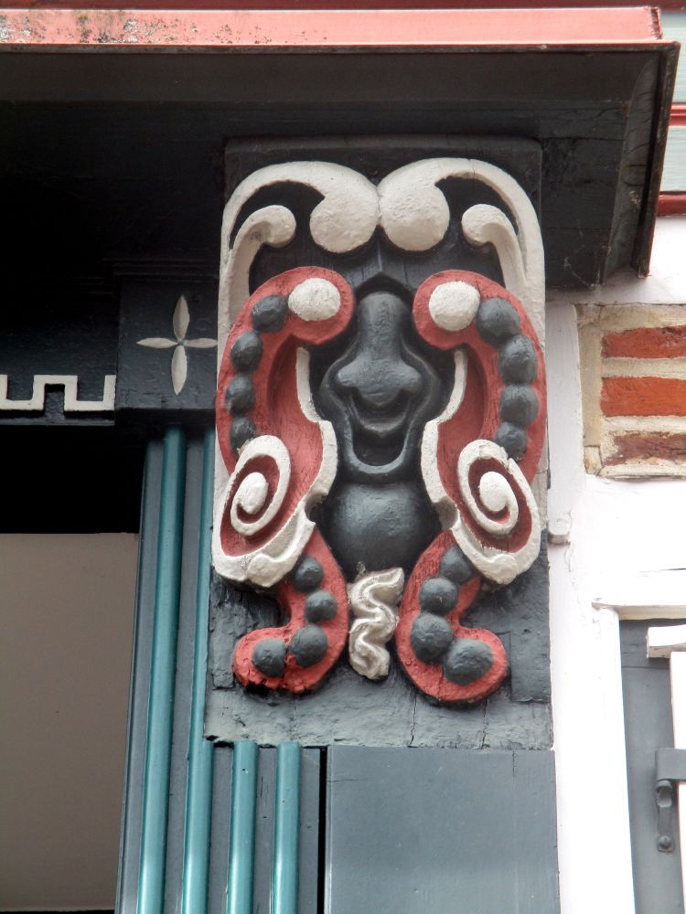 Eine Schnitzerei an einem Hausbalken. Eine fröhliche Maske die Schwarz, weis, Rot bemalt ist.