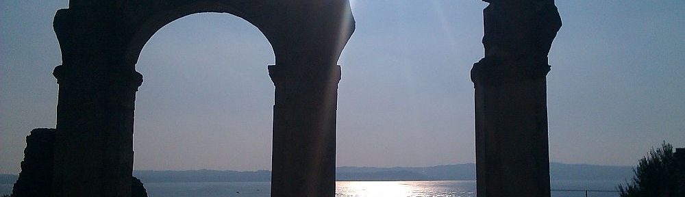 Die Überreste der Ruinen an den Grotten des Catull. Zu sehen ist ein Säulenbogen und eine danebenstehende beschädigte Säule. Sie zeichnen sich als Schatten vor dem Wasser des Gardasees und einem blauen Himmel ab.