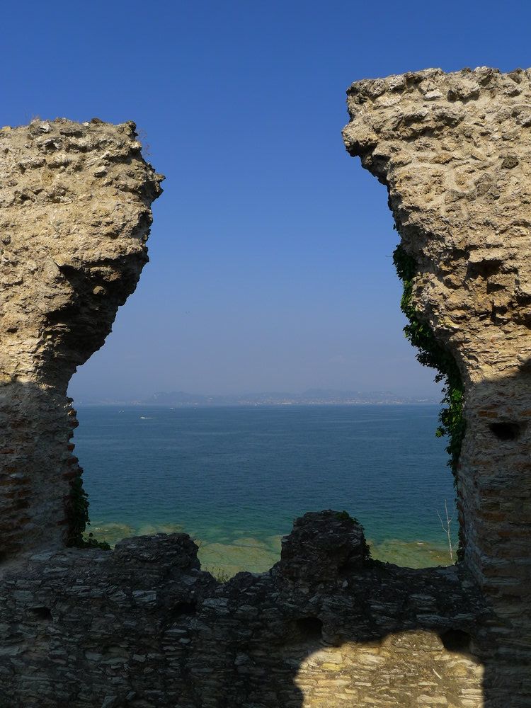 Blick durch die Ruinen der Grotten des Catull hindurch auf den Gardasee. Im Vordergrund sind links und rechts Ziegelmauern, dahinter liegt grünblaues Seewasser, und ein blauer Horizont.