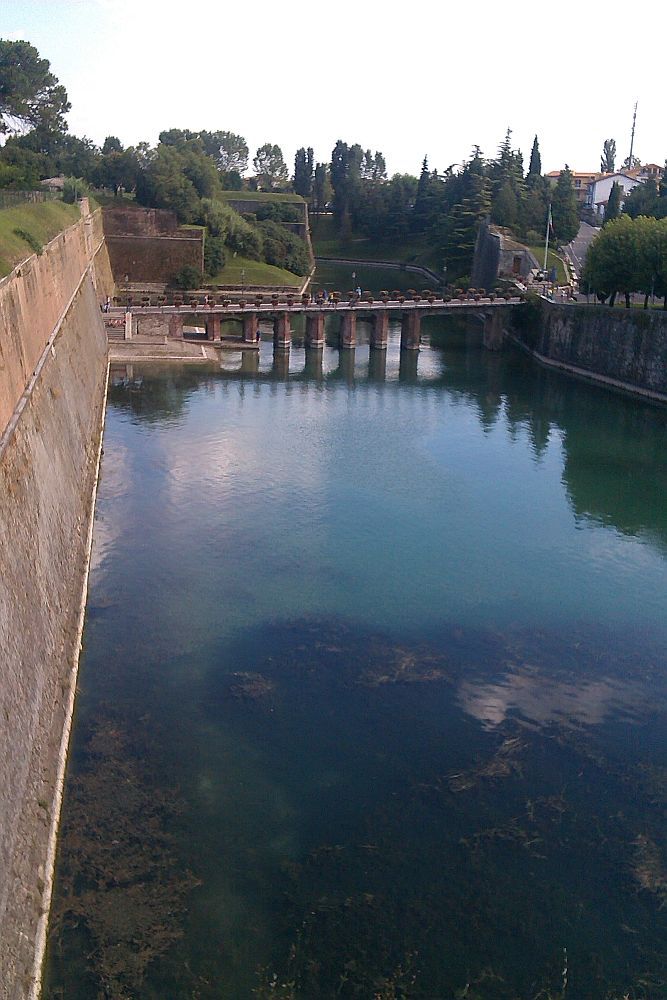 ine Brücke aus roten Zigeln verläuft übr den Blauen Mincino. Die Brücke ist mit roten Blumen Gschmückt. Deutlich zu sehen ist, das von zwi Bastionen einsehbar ist wer die Brücke überquert um zu der Festung zu gelangen.