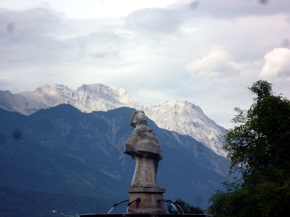 Ein Brunnen aus Granit mit einer Brunnenfigur in der Mitte. Ein Mann aus Granit, der in den Himmel schaut. Alles steht vor einem Alpenpanaorama.