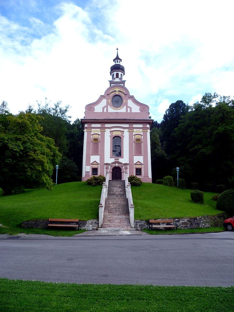 Eine Kapelle mit geschwungenen Rokokogiebel von Vorne. Die Kapelle ist weiß, mit hellroten Schnörkeln. Sie hat einen Turm und liegt auf einem grün bewachsenen Hügel.