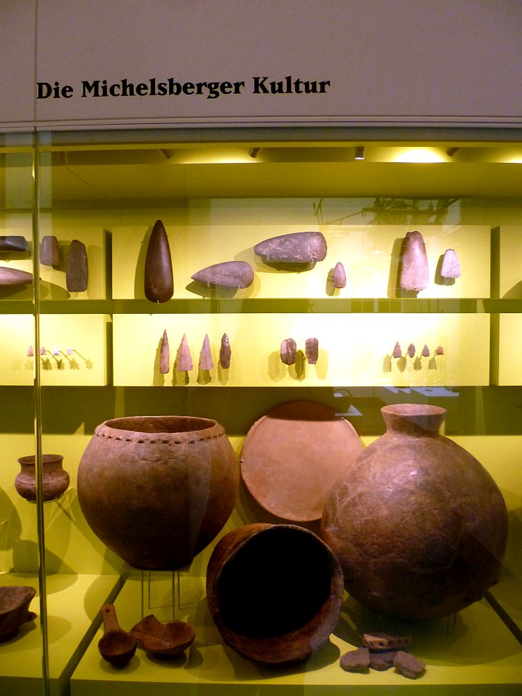 Eine Aufstellung von Funden der Michelsberger Kultur in einer Vitrine. Die Vitrine ist angeordnet wie eine Typologietafel. Große Steingeräte oben, darunter kleinere und auch Pfeilspitzen, darunter Keramik. Riesige runde Becher.