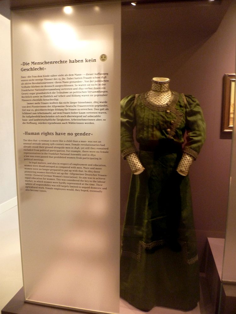 Eine Vitrine mit der Aufschrift, "Menschenrechte haben kein Geschlecht". In der Vitrine ist ein dunkelgrünes knöchellanges Kleid mit Puffärmeln.