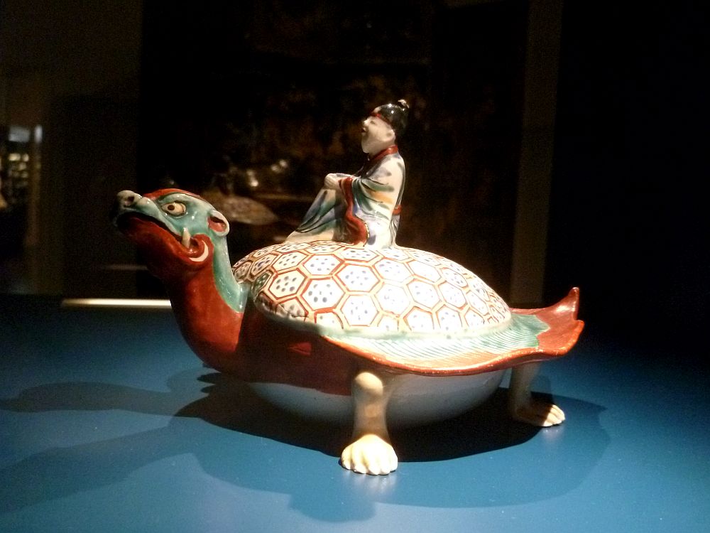Ein Mann reitet auf einer Schildkröte. Er trägt einen Kimono. Die Schildkröte ist Blau und Grün. Es handelt sich um eine Porzellanfigur.