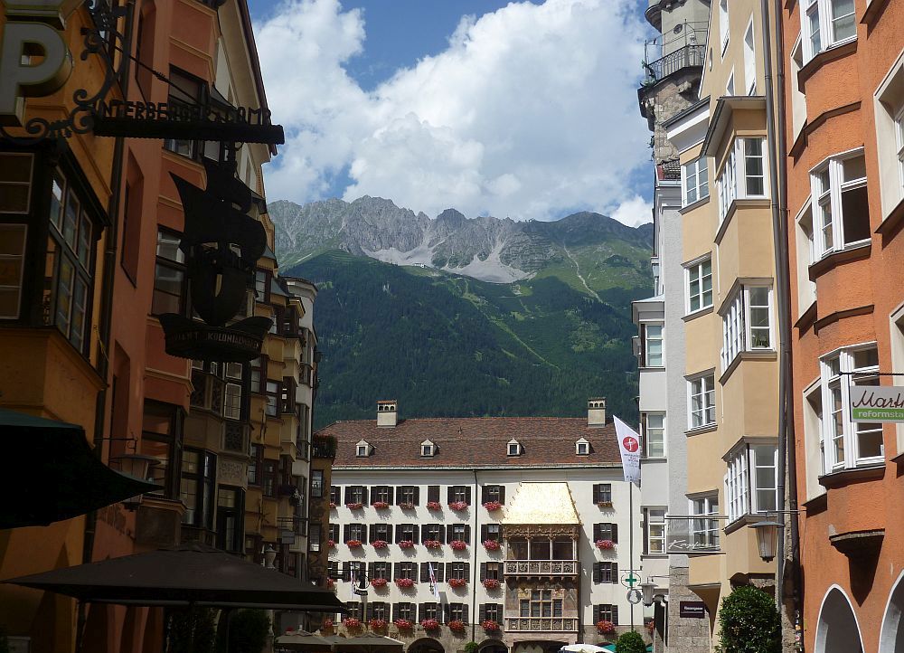Das goldene Dachl am Ende eines Marktplatzes. Im Vordergrund links und rechts alte Erkerhäuser, in der Mitte das Haus mit dem goldenen Dach und am Horizont die gigantischen Alpen in Grün bewachsen.