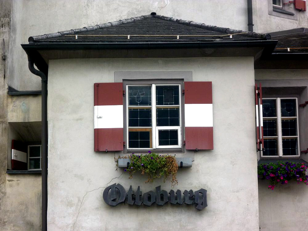 Ein Erker aus grauem Stein. Es gibt ein Fenster mit weiß-roten Fensterläden. Darunter ein Schild mit dem Namen "Ottoburg"