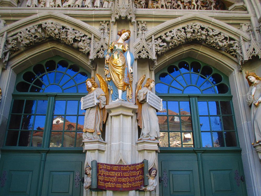 Bemalte Sandsteinfigur auf einem Sockel vor zwei Spitzbögentüren. Die Figur zeigt Justizia, trägt ein blaues Kleid mit einer Art goldenen Toga und goldenen Applikationen. In der rechten Hand hält sie ein schwer leicht erhoben, in der Linken eine goldene Wage, am lang hängenden Arm. Die Figur ist links und rechts durch zwei Sandsteinengel gerahmt, die etwas niedrieger stehen. Sie tragen ein beiges Kleid und haben goldenes Haar, und goldene Flügel.