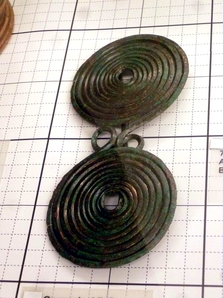 Eine Brillenfibel auf einem karierten Hintergrund. Sie besteht aus einem Draht, der zu zwei gegenläufigen Spiralen gewickelt wurde. Die Fibel ist ca. 15 cm lang.