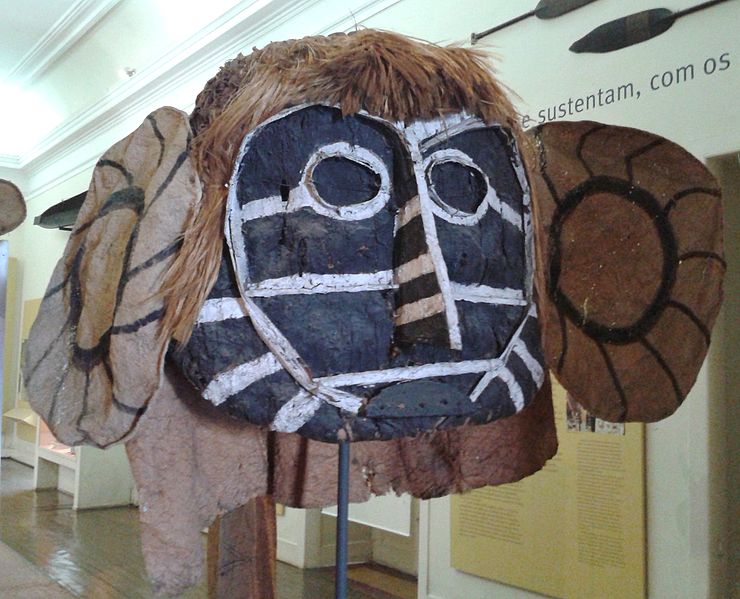 Eine große Maske aus Holz und Leinenstoff. Das Gesicht ist aus Holz gefertiegt. Es ist schwarz und mit weißen Streifen bemalt. Haare und Ohren bestehen aus braunem Stoff.