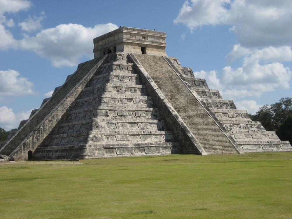 Die Pyramide von Chicen Itza. Ein spitz zulaufender Bau aus grauem Stein. Er ist in Stufen angelegt und hat große Treppen, die nach oben zu einem Altar führen.