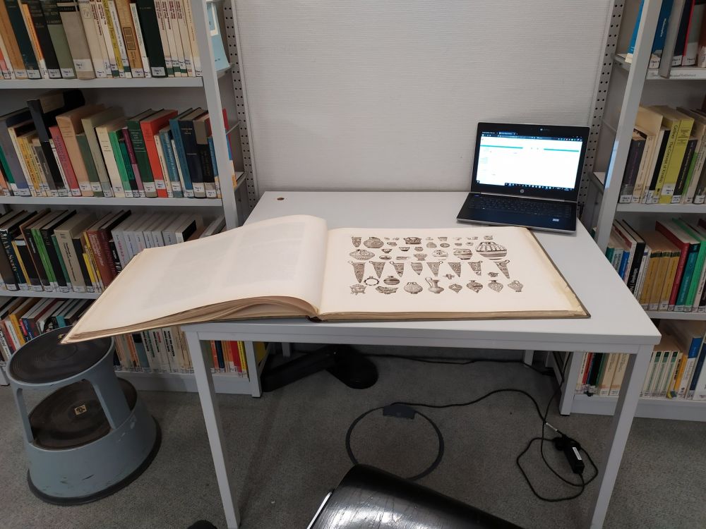 Auf einem Tisch zwischen zwei Bücherregalen steht ein Laptop und ein Buch liegt auf dem Tisch. Das Buch ist gut doppelt so groß wie der Tisch.