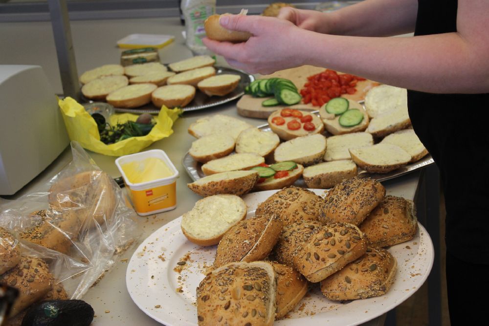 Zwei Hände sind zu sehen, die ein Brötchen schmieren. Im Hintergrund liegen viele geschmierte Brötchen.