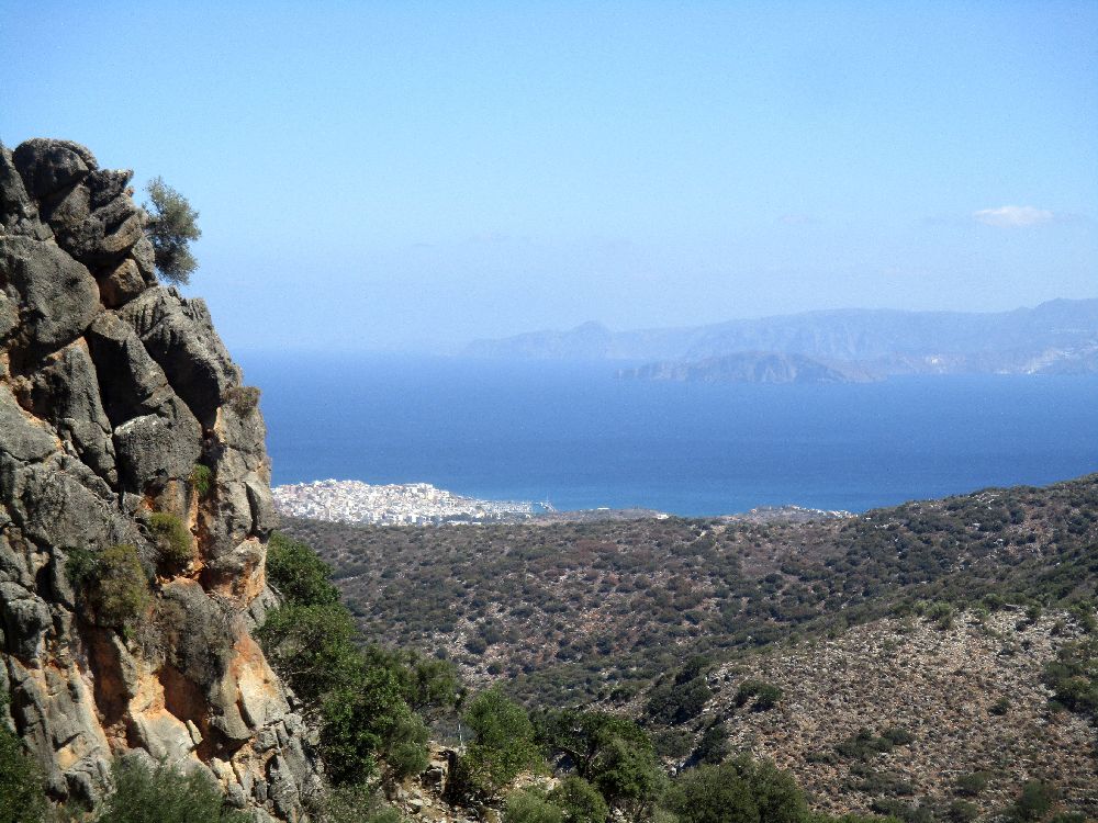 Die Aussicht von einem Berg aus. Links sind noch ein Paar Felsen zu sehen, im Vordergrund ersteckt sich eine Landschaft. Eine Stadt, die aus weißen Häusern gebaut ist zieht sich ins Meer, das sich dahinter weithin sichtbar ausbreitet, und ganz im hintergrund ist noch ine Bergkette zu erkennen, von der anderen Seite der Meeresbucht.