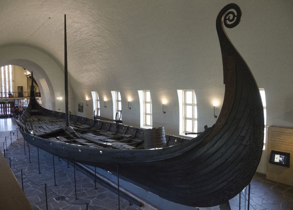 Das Osebergschiff so wie es heute Im Museum Steht. Es bsteht aus planken, und hat einen hoch gezogenen Bug, der in einem Kringel endet.