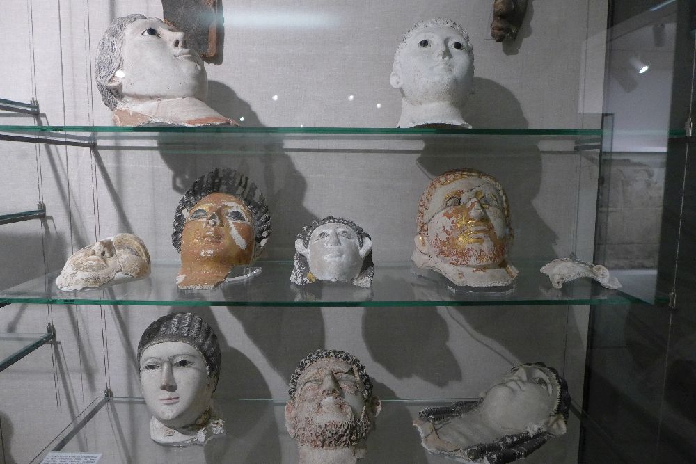 Vitrine mit 8 Kartonagemasken. Eine ist vergoldet, zwei sind braun bemalt, die anderen weiß.