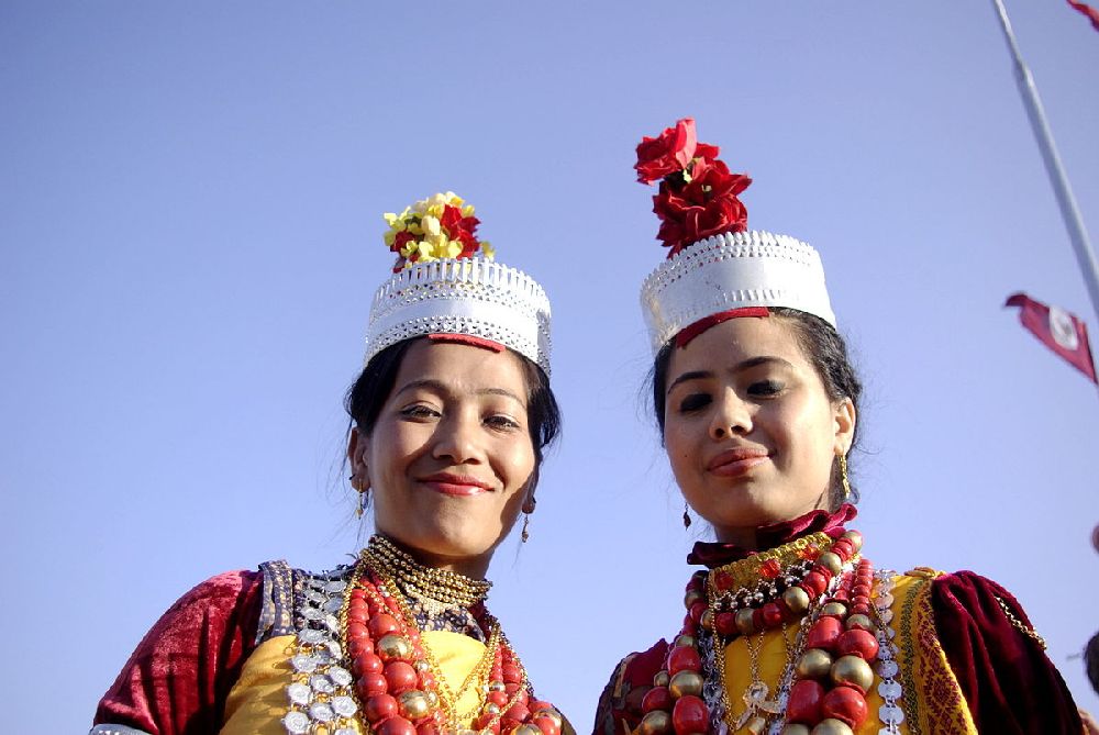 Zwei Khasi Mädchen. Sie Tragen silberne Kronen mit gelben und Roten Federbüschen oben drauf. Ihre Kleidung besteht aus gelben und roten Farben, sie ist allerdings kaum zu sehen, da die Mädchen Massiv Ketten tragen, die ebenfalls Silbern gelb und rot sind