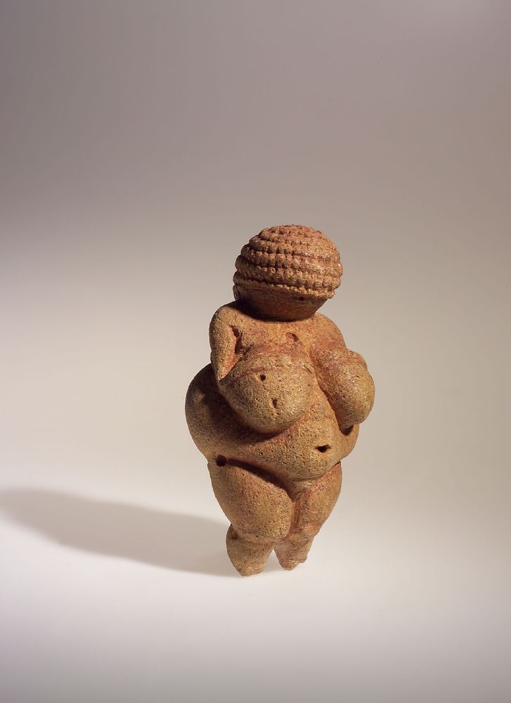 Die Veneus von Willendorf von rchts. Eine shr Dicke stilistisch Dargestellte Frau mit iner Kurzhaar oder Felchtfriesur. Evtl. trägt sie auch eine Haube. Die Frau hat Dünne Arme, die sie auf ihre Brüste gelegt hat. Diese sind sehr Dick, und auch der Bauch und die Hüften sind sehr füllig.