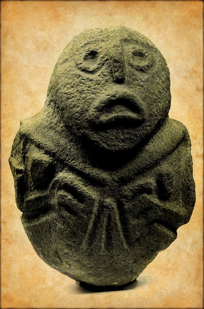 Eine Kopfskulptur mit hervor gearbeiteter Vulva. Die Darstellung des Gesichts erinnert an einen Karpfen.