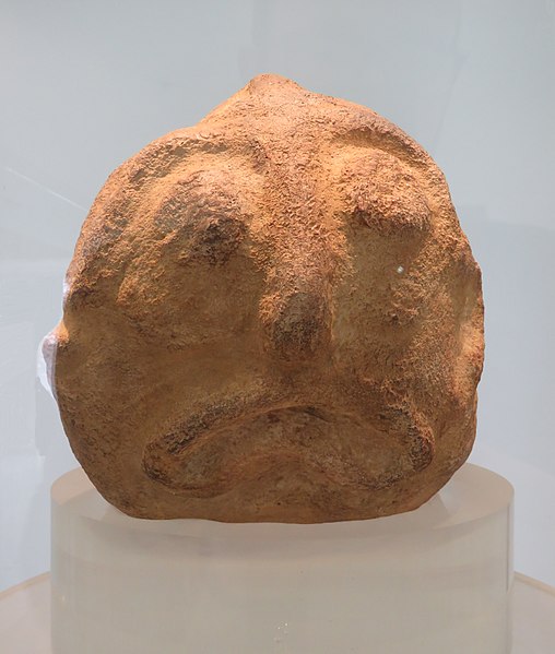 Ein Terrakottafarbener Stein. Er ist rund, und zwei kugelrunde Augen, eine Nase und ein schlauchartiger nach unten gebogener Fischmund sind in den Stein graviert.