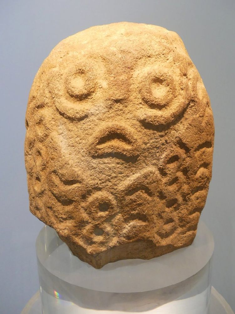 Eine Figur aus Lepenski Vir. Sie hat viele Ornamente und in der Mitte ein Gesicht. Die Augen sind kreisrund, und der Mund ist Schlauchartig und mies gelaunt nach unten gebogen.