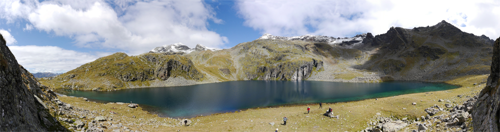 Der Alkuhuser See. Das Wasser ist tif Blau, die Umgebung ist gelbgrün bewachsen, und im Hintergrund erbeben sich Grüne bewachsene Berggipfel.