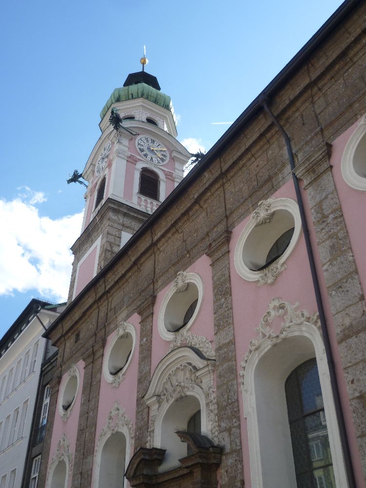 Die Spitalskriche von Innsbruck aus extremer Froschperspektive. Die Kirche ist Rosa gestrichen. Eine Reihe runder Fenster ist zu sehen. Und ein Turm mit einem grünen Zwiebeldach aus Kupfer.