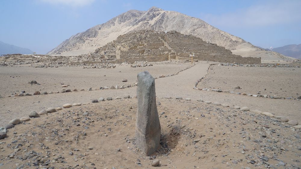 Im Vordergrund ist eine Große Steinsäule zu sehen, und im hintergrund führt ein mein Steinen begrenzter Weg zu einer Pyramide.