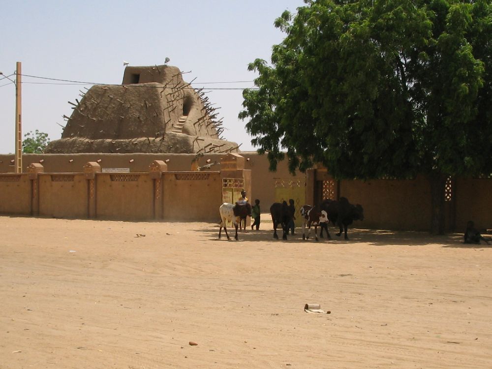 Eine Pyramide aus Lehm hinter einer Mauer. Im Vrdergrund treibt ein Junge Rinder über die Strasse.