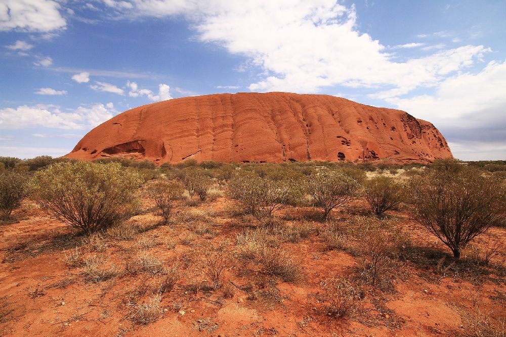 der Uluṟu, ein Roter Berg umgebe von einer Steppe. Deer Berg ist geformt wie ein langes flaches oval. Er ist Oben sehr flach.