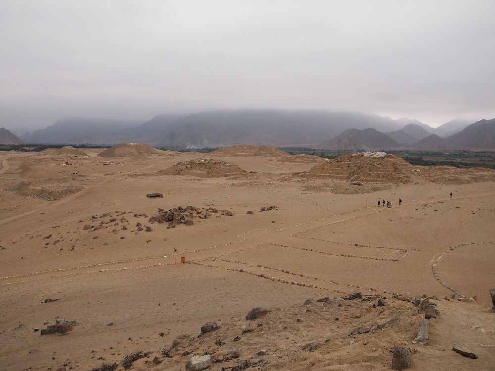 Im Hintergrund verschwinden die Anden in den Wolken. Im Forderung dind die Ruinen von Caral zu sehen. Eine Strasse führt in Richtung von Pyramieden, davorgelagert ist ein bereich mit vielen kleinen Gebäuden und eine Prachtstrasse führt aus dem Bild heraus.