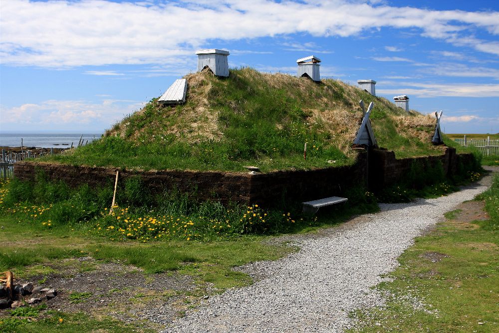 Ein Haus aus L´Angse aux Medows. Es handelt sich um einen Holzbau der rundum mit Gras bewachsen ist. Alles ist sehr Grün. Löwenzah Blüht an dem Haus und der Himmel ist Blau.