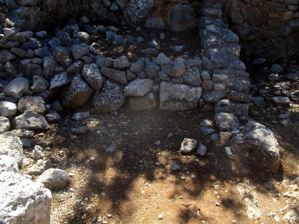 Ein Raum aus grauen Stein gemauert auf gelben Sand. Es scheint eine gemeuerte empore oder einen Altar gegeben zu haben. 