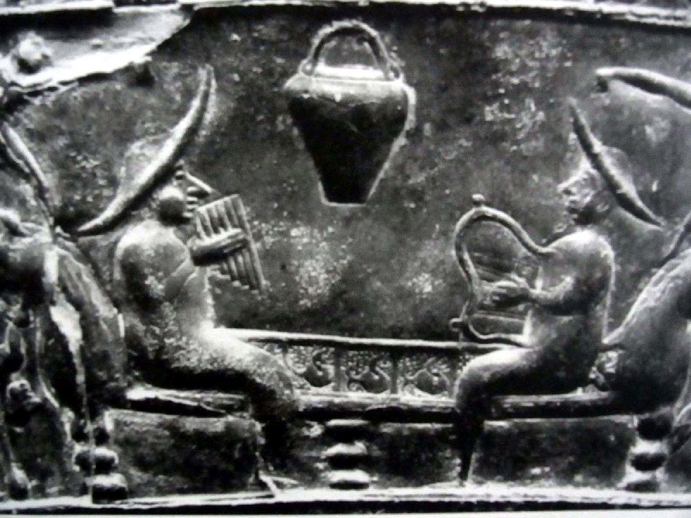 Eine Szene der Bronzesitula Bolonga Certosa. Das Relief zeit zwei Männer die sich auf einer Bank eggenüber sitzen, der Mann zur rechten spiel Panflöte, der zur linken spielt eine Leier. Beide Männert tragen riesiege Sonnenhüte.