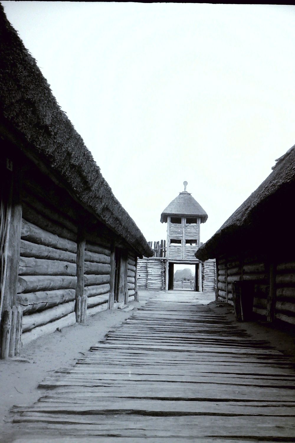 Das Eingangstor von innen in schwarzweißfotografie. Links und recht befinden sich Reihenhäuser, in der Mitte ein Bohlenweg und am Ende ein riesieges Tor