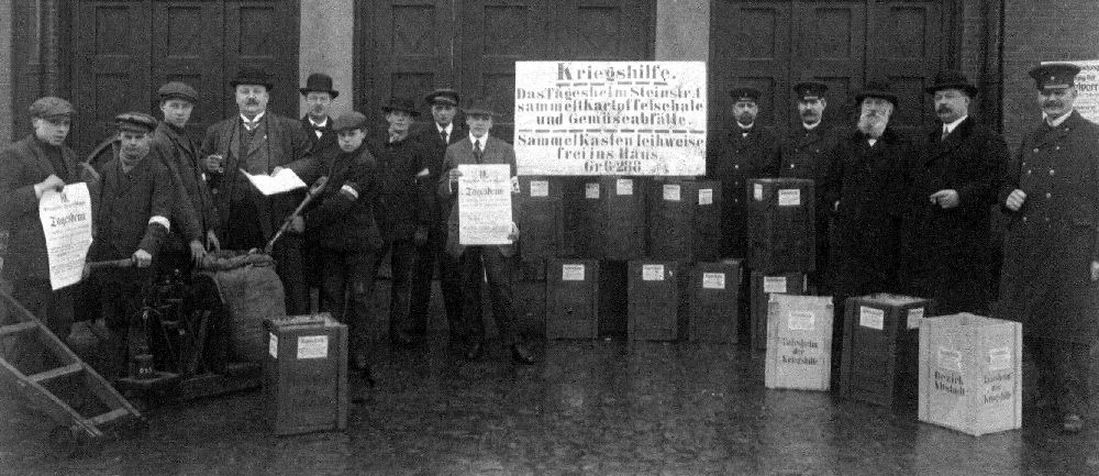 Ein Duzend Männer stehen um ein Schild herum, mit der Aufschrift "Sammelt Kartoffelschalen" Sie sammeln für eine Notstelle in der Steinstr. in Hamburg.