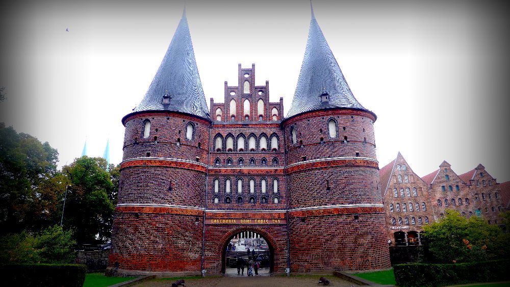 Das Holstentor im Regen. Der Himmel ist grau, und das Tor mit den zwei Türmen leuchtet Rot vor der Stadt Lübeck.