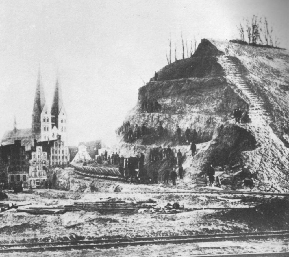 Ein historisches Foto. Im Vordergrund ein Wall, der drei mal so hoch ist wie die dahinterliegende Kirche, Menschen über mehrere Stockwerke im Wall von Lübeck, die diesen Abtragen.