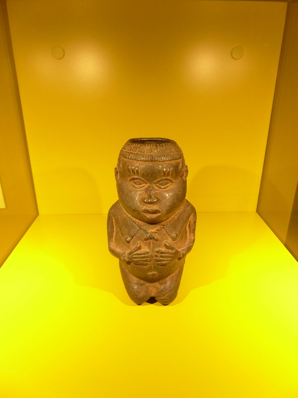 Ein Bronzegefäß in Form einer Schwangeren Frau. Die Frau hat ein Loch im Kopf, da es sich um ein Salbgefäß handelt.