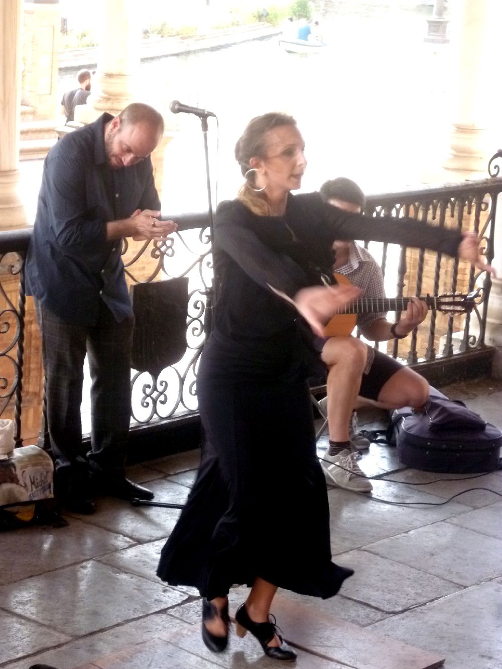 Eine Flamencotänzerin, sie Tanzt, hat beide Arme nach Vorne Ausgestreckt dabei. Im Hintergrund klatscht ein Mann, ein zweier Spielt dazu sitzend Gitarre.