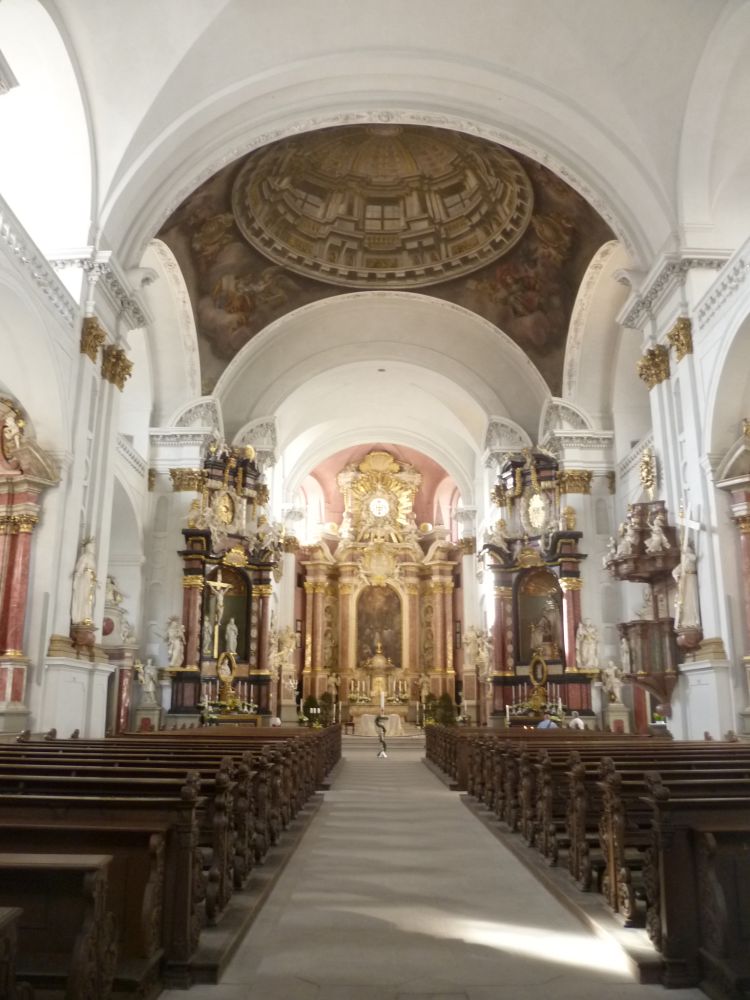 Eine helle schlicht gestalltete Barockkirche von innen. Holzbänke, und weiße Wände.