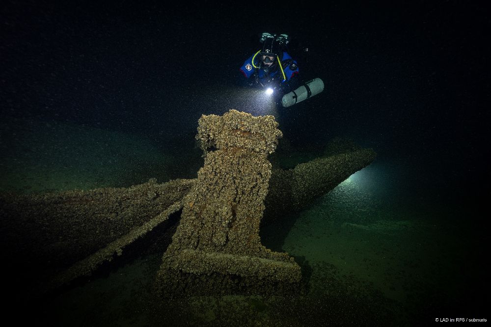 Taucher oder Taucherin im tiefdunklen Wasser. Die Person leuchtet mit einer Lampe ein mit Algen überwachsenes Schiffswrack an.