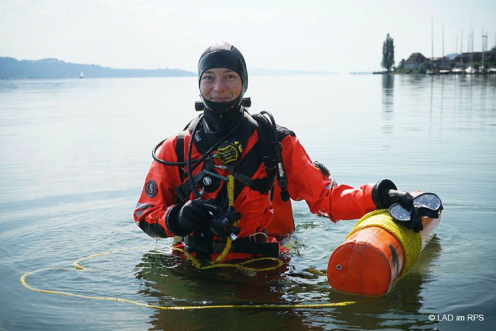 Eine Frau in einem roten Taucheranzug, die bis zur Hüfte im Bodensee steht.