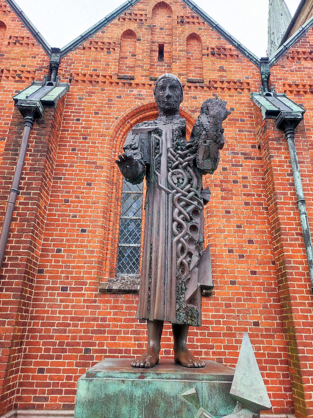 Eine Bronzeskulptur vor einer Backsteinwand. Die Skulptur zeit einen Hochgewachsenen glatzköpfigen Mann. Eine Hälfe von ihm besteht aus einer Art Knotenornament.