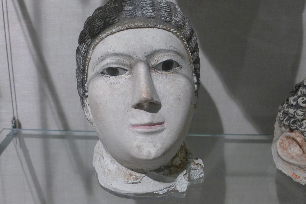 Eine weiße Maske mit schwarzem Haar. Die Frau hat eine lange große Nase, schmale roséfarbene Lippen und schmale Augen mit Glaseinlage.