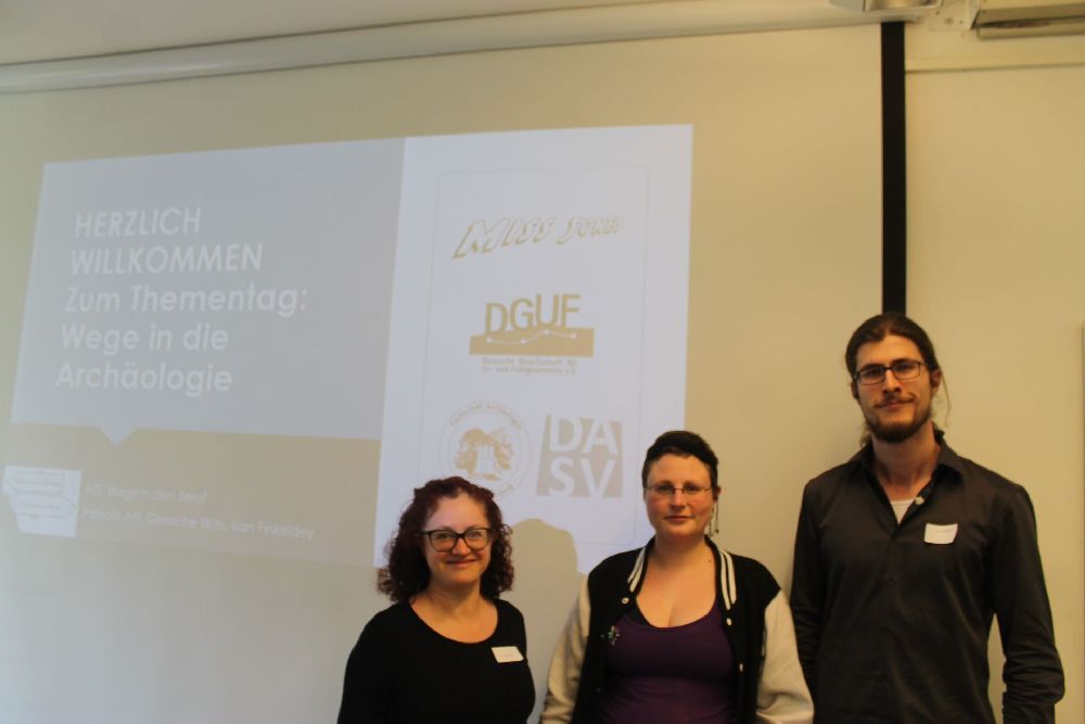 Drei Personen stehen vor einer Beamerwand, auf der Zahlreiche archäologische Organisationen ihre Kürzel hinterlassen haben. Es handelt sich um Patricia Arlt, Geesche Wilts und ilian Finkeldey.